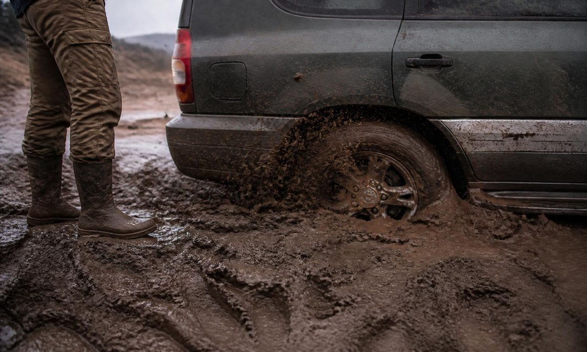 A crossover with a rear wheel buried and spinning in deep brown mud as a person in boots looks on